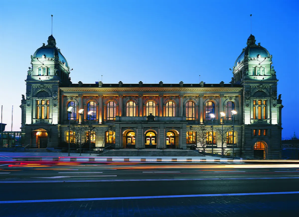 Die Historische Stadthalle Wuppertal bei Abenddämmerung, beleuchtet mit warmem Licht, aufgenommen von der gegenüberliegenden Straßenseite.